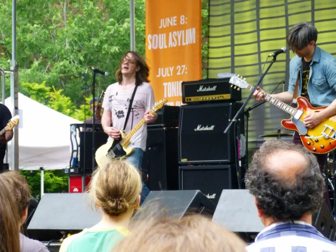 This image is clicked in a concert. There are two men playing guitar. Behind them there are...
