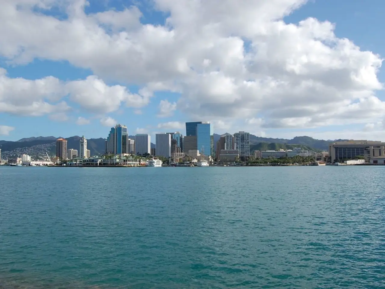 In this picture we can see water. In the background of the image we can see buildings, trees, hills...