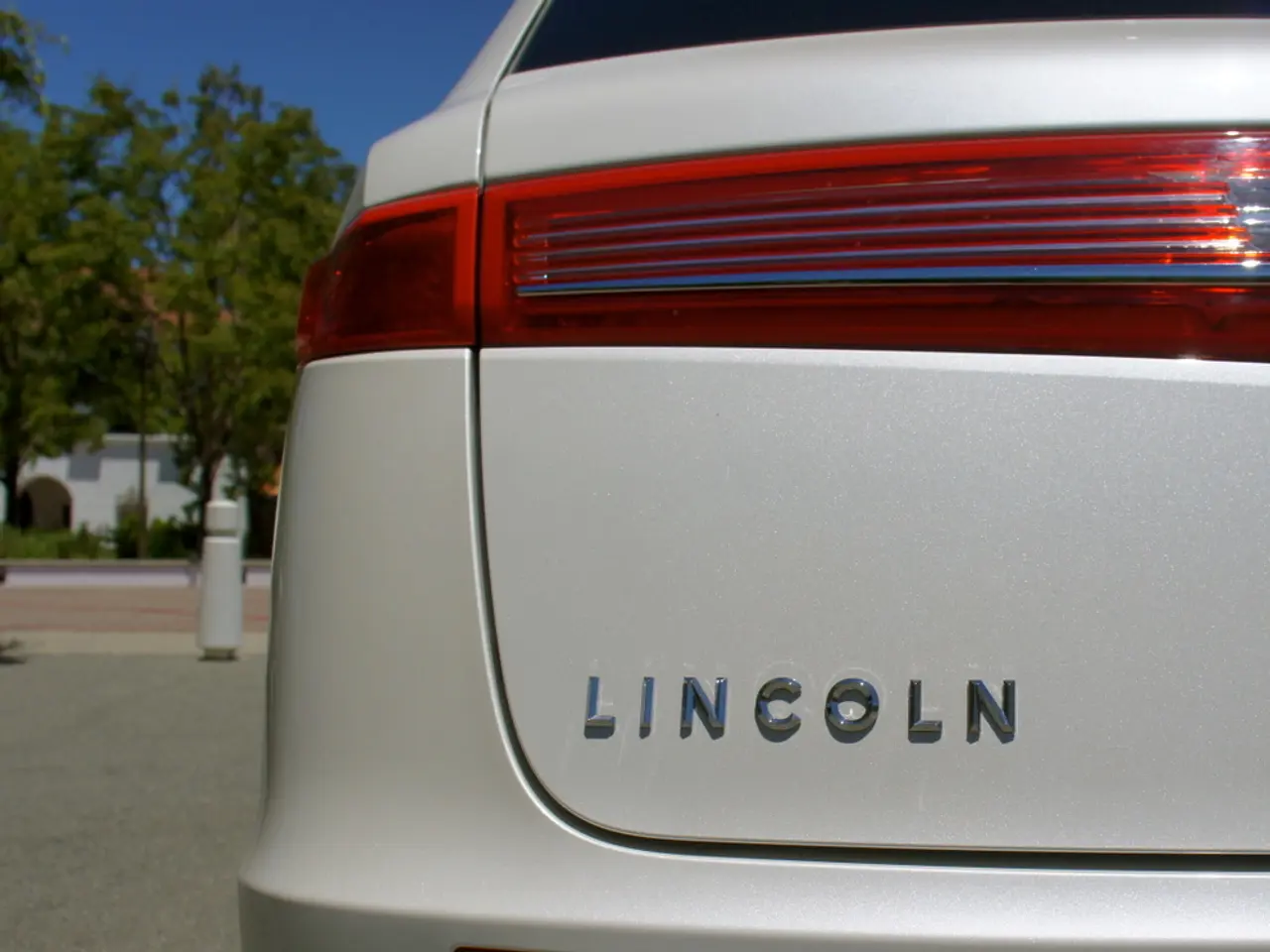 In this image there is a car beside that there are some fencing poles, trees and buildings.