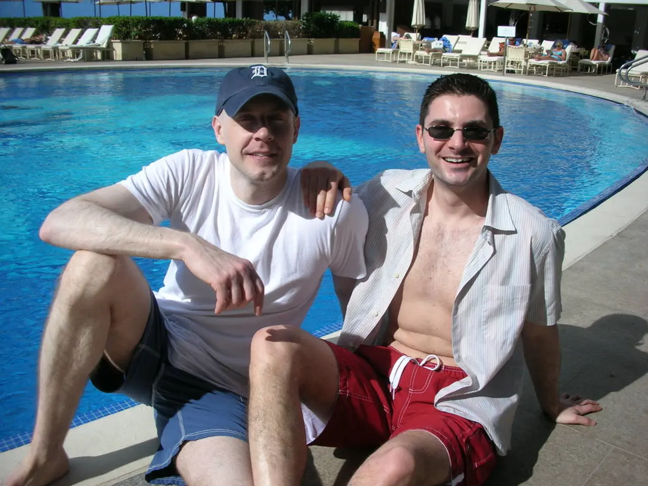 In the image there is a swimming pool beside it there are two men sitting and they are smiling. One...