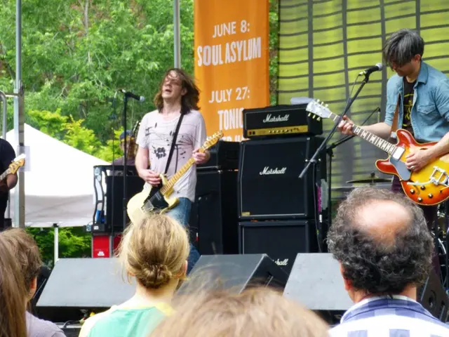 This image is clicked in a concert. There are two men playing guitar. Behind them there are...