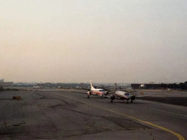 This picture shows a couple of aeroplanes on the runway and we see buildings and trees and we see a...